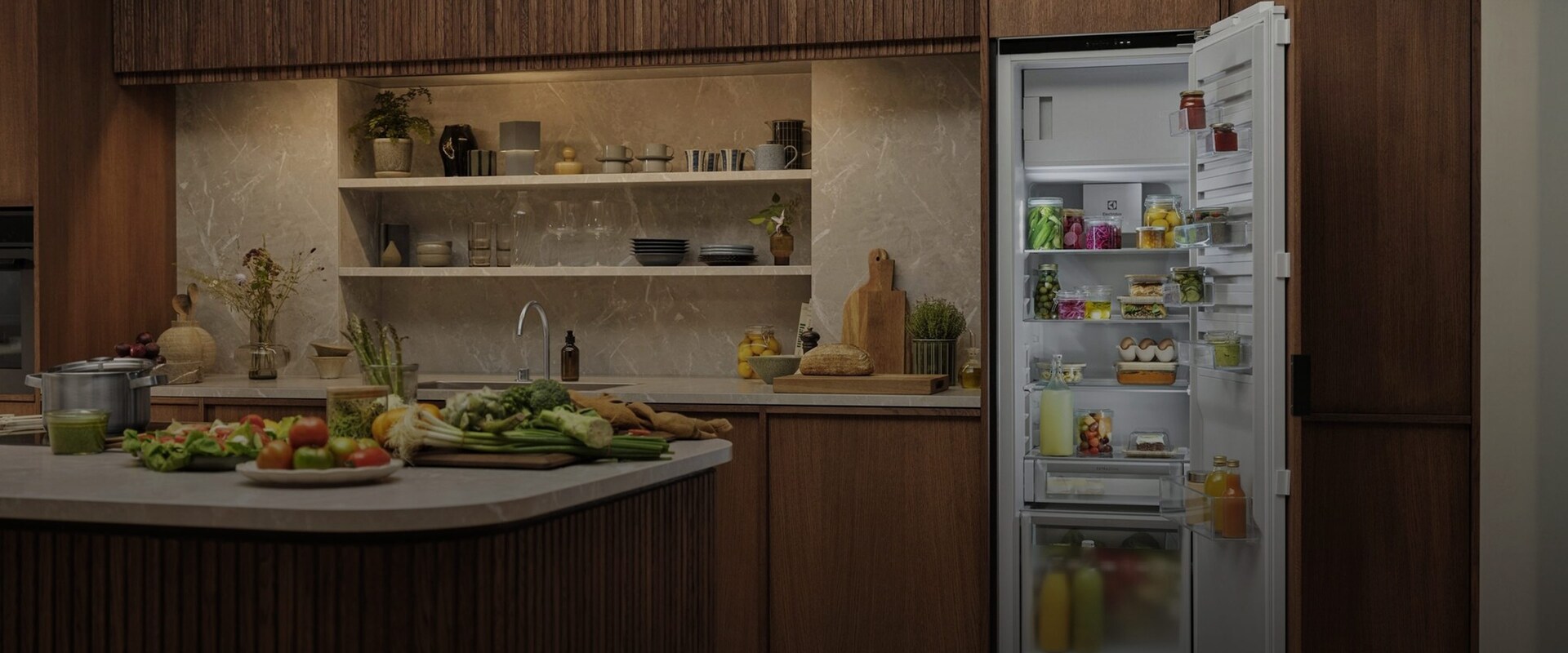 Yleiskatsaus avonaiseen, modernissa keittiössä sijaitsevaan integroituun Electroluxin jääkaappiin, joka on täytetty tuoreilla raaka-aineilla, pulloilla ja säilytysrasioilla.